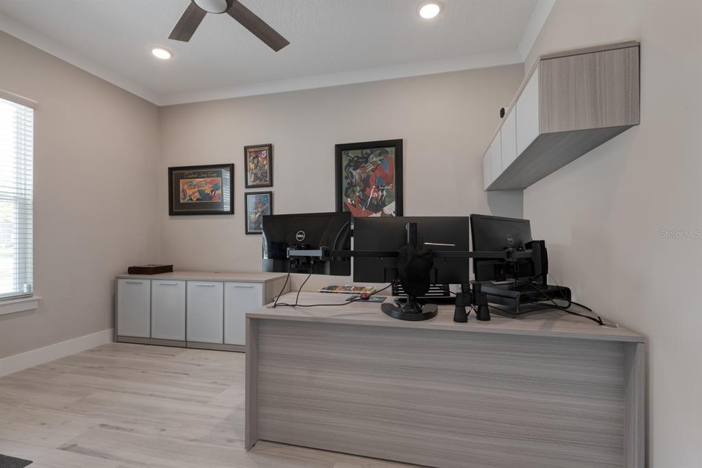 A home office features a large desk with multiple monitors, wall cabinets, and light wood flooring — 25962 HIGH HAMPTON CIRCLE, SORRENTO, FL