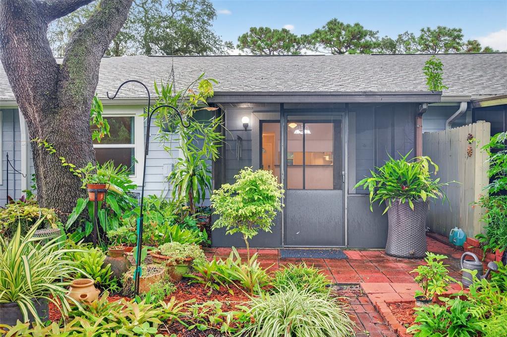 A brick patio leads to a screened-in entryway surrounded by lush potted plants and trees — 39650 US HIGHWAY 19 N UNIT 565, TARPON SPRINGS, FL