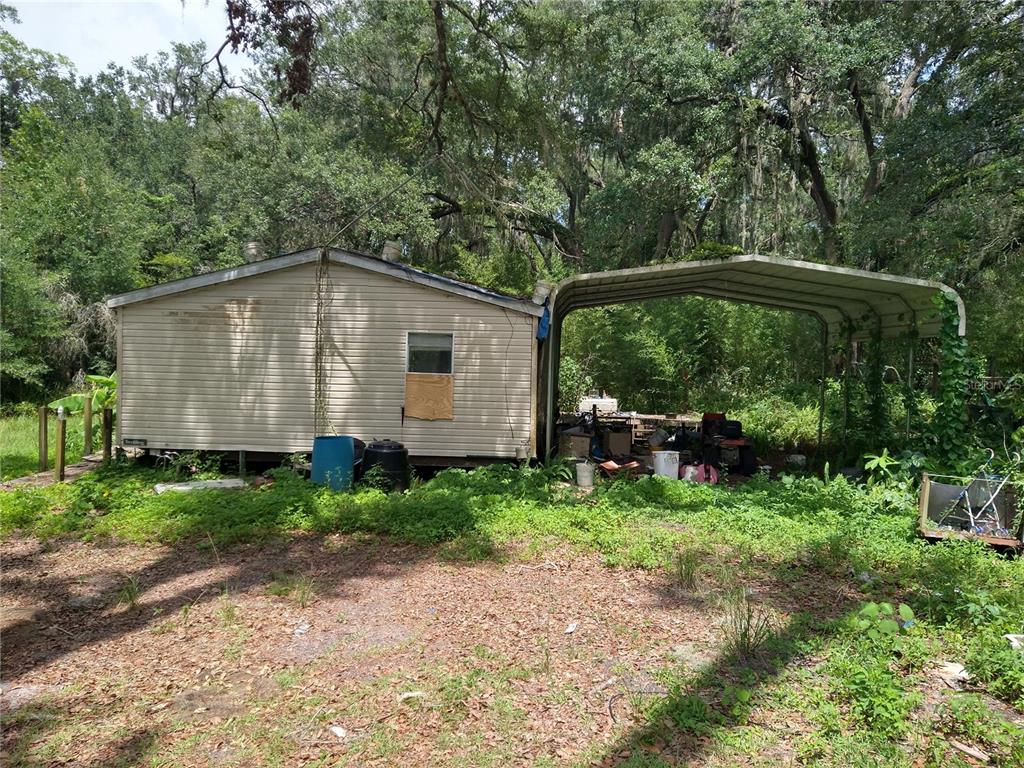 A single-story mobile home with an attached metal carport sits on a wooded lot — 10616 SHADY DRIVE, HUDSON, FL