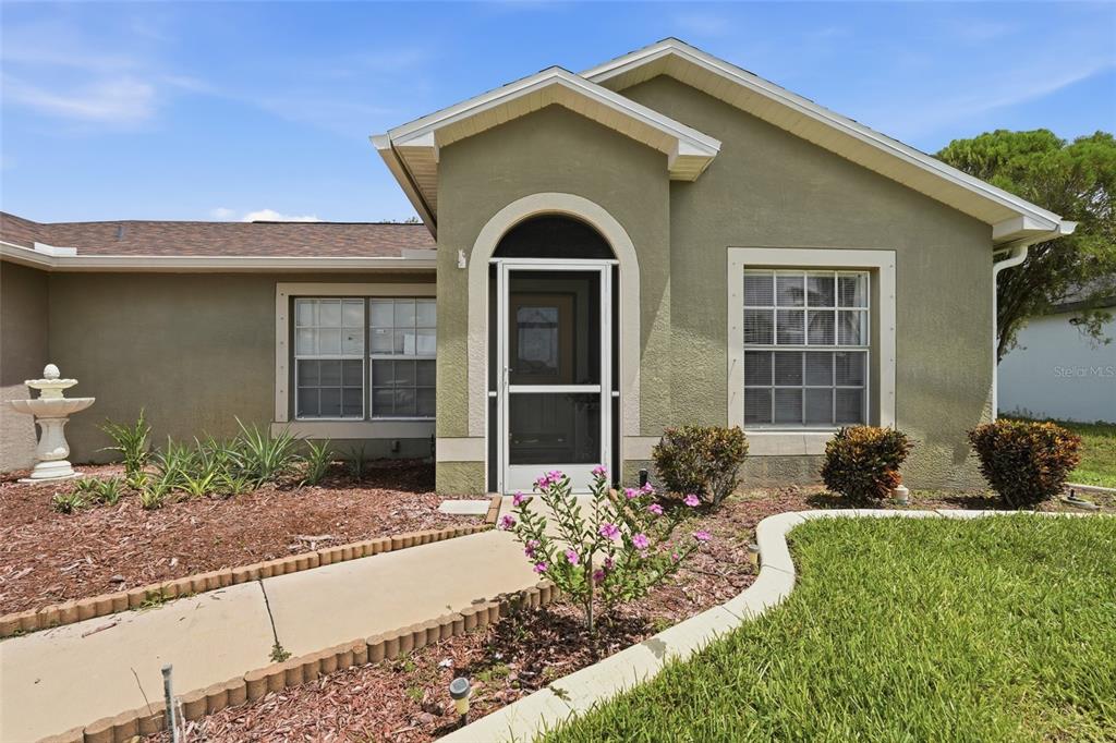 The exterior of a single-story house features a green stucco facade, arched entryway, and landscaping — 4112 10TH STREET W, LEHIGH ACRES, FL