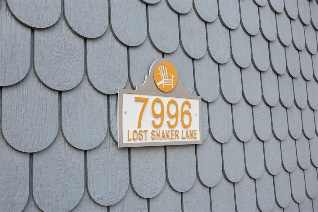 A house number sign is mounted on a wall with gray, rounded shingle siding — 7996 LOST SHAKER LANE, KISSIMMEE, FL