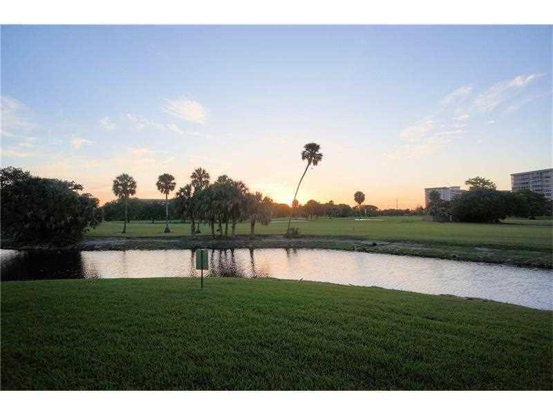 A grassy lawn overlooks a small pond with palm trees and buildings in the distance — 565 Oaks Lane # 103, Pompano Beach FL 33069, POMPANO BEACH, FL