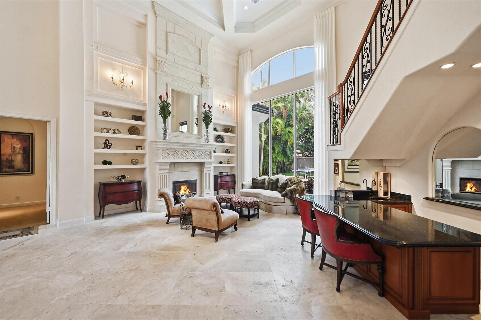A high-ceilinged living room features a stone fireplace, built-in shelves, a bar, and large windows — 2425 NW 49th Lane, Boca Raton FL 33431, BOCA RATON, FL