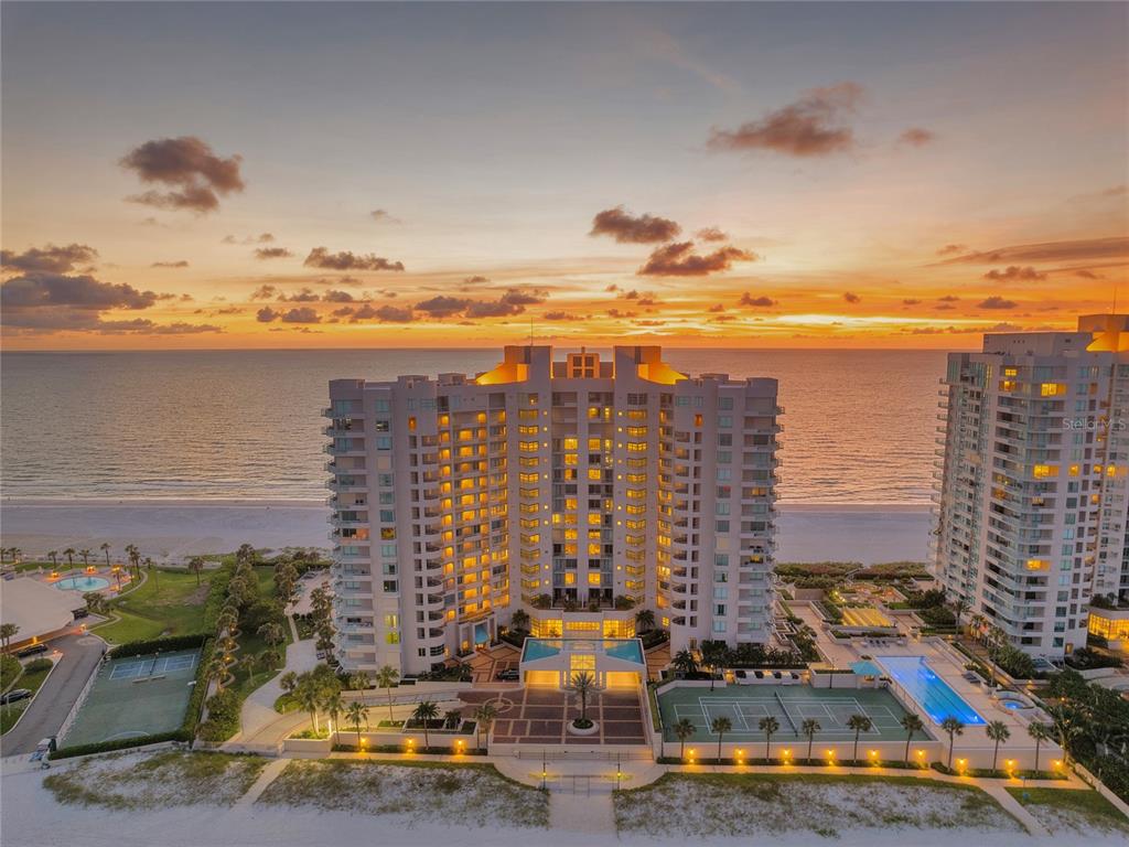 An aerial view of a beachfront high-rise condominium building at sunset with tennis courts — 1560 GULF BOULEVARD UNIT 0000, CLEARWATER BEACH, FL