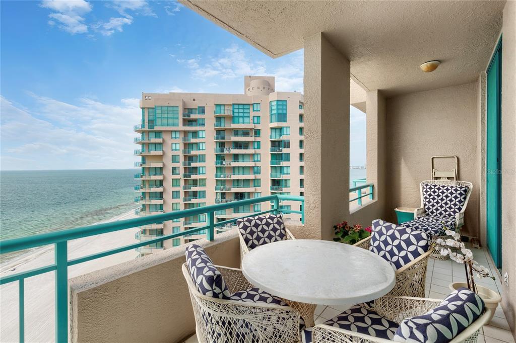 A balcony with a round table and chairs overlooks the beach and a neighboring building — 1560 GULF BOULEVARD UNIT 0000, CLEARWATER BEACH, FL