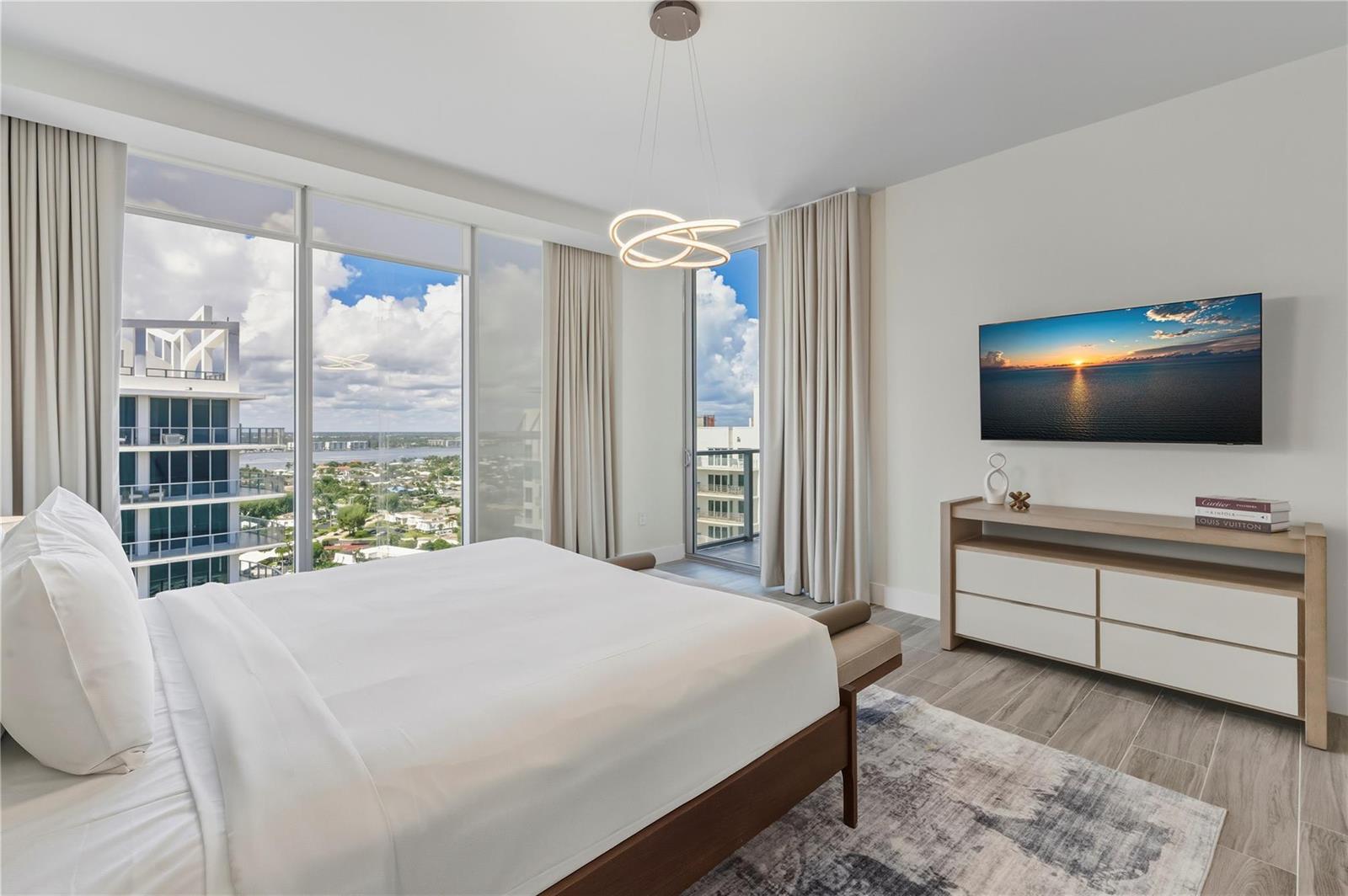 A modern bedroom features a bed, a wall-mounted television, and floor-to-ceiling windows with city views — 3100 N Ocean Drive # P1802, Riviera Beach FL 33404, RIVIERA BEACH, FL