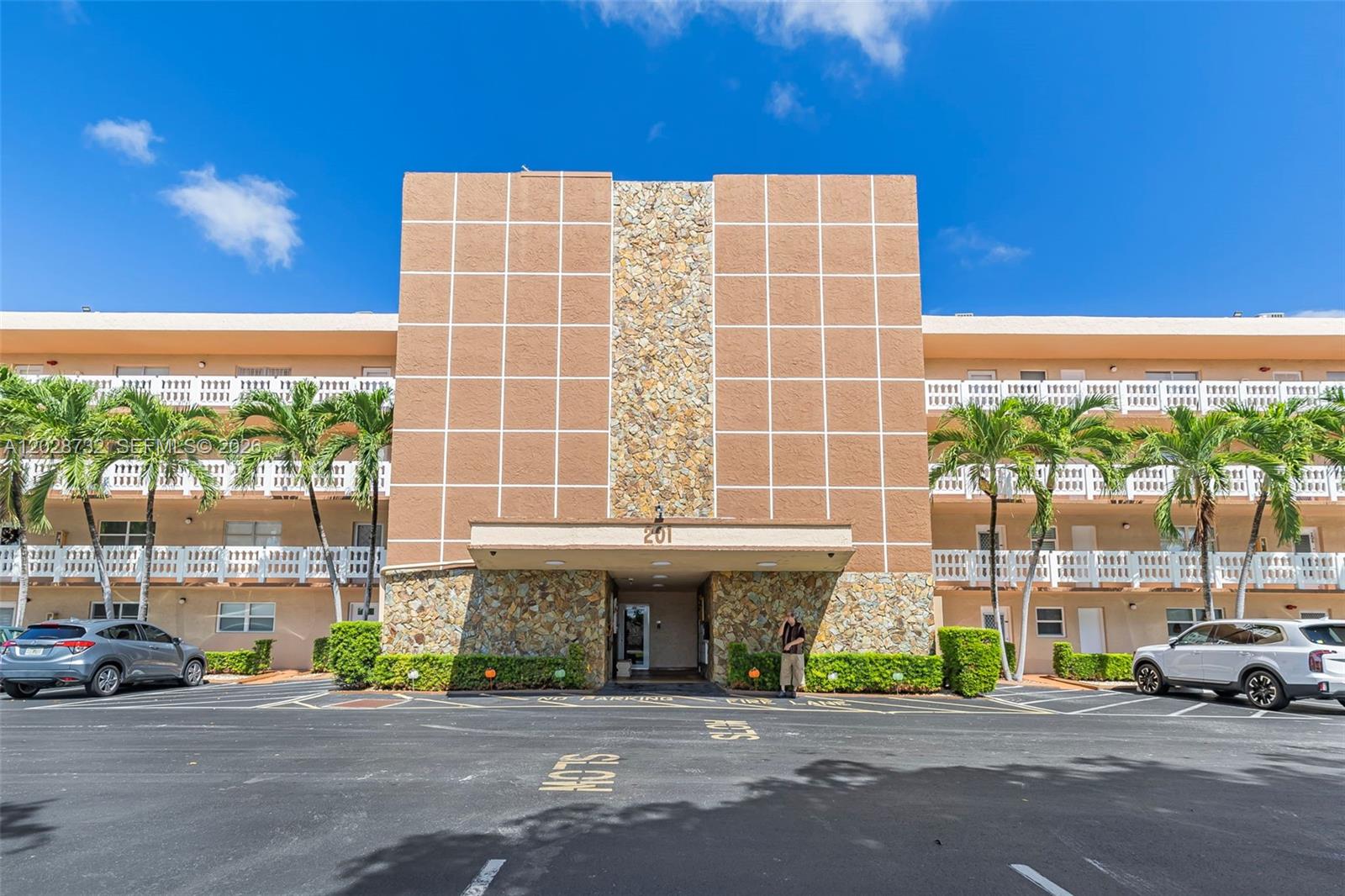 The exterior of a multi-story apartment building features a stone-accented entrance and parking lot — 201 SE 11th Ter # 102, Dania Beach FL 33004, DANIA BEACH, FL