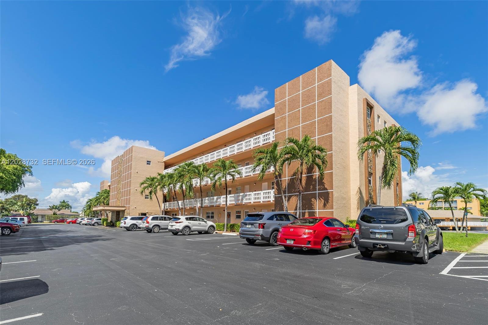 A multi-story apartment building with a parking lot and palm trees under a blue sky — 201 SE 11th Ter # 102, Dania Beach FL 33004, DANIA BEACH, FL