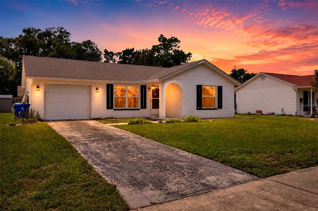 A single-story white house with a garage and driveway sits under a vibrant sunset sky — 7412 HUMBOLDT AVENUE, NEW PORT RICHEY, FL