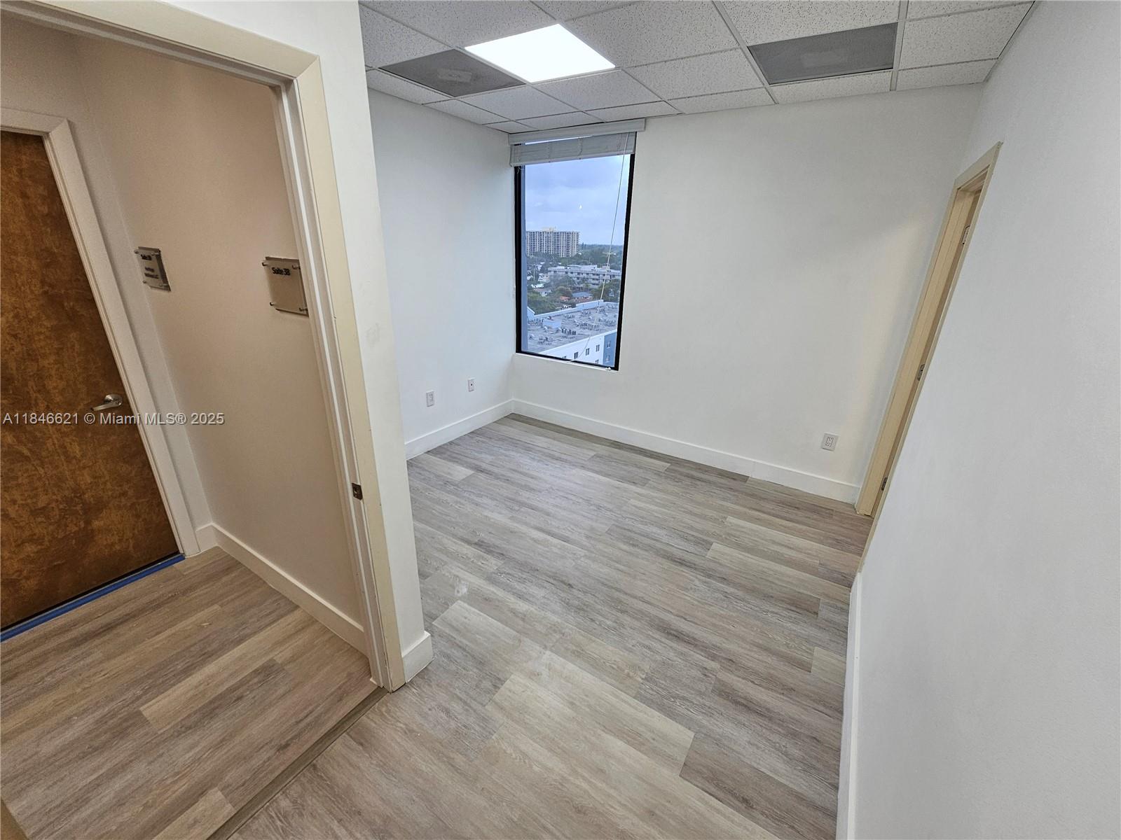 An empty office space features light wood-look flooring, white walls, and a window with views — 12550 Biscayne Blvd # 11, North Miami FL 33181, NORTH MIAMI, FL