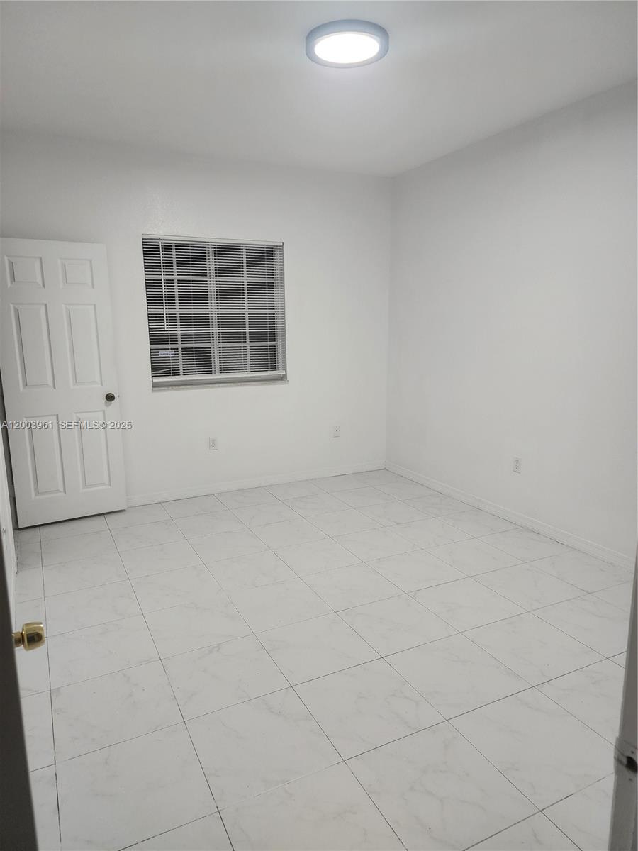 An empty room features white walls, white tiled flooring, a window with blinds, and a door — 6972 NW 179th St # 4, Hialeah FL 33015, HIALEAH, FL