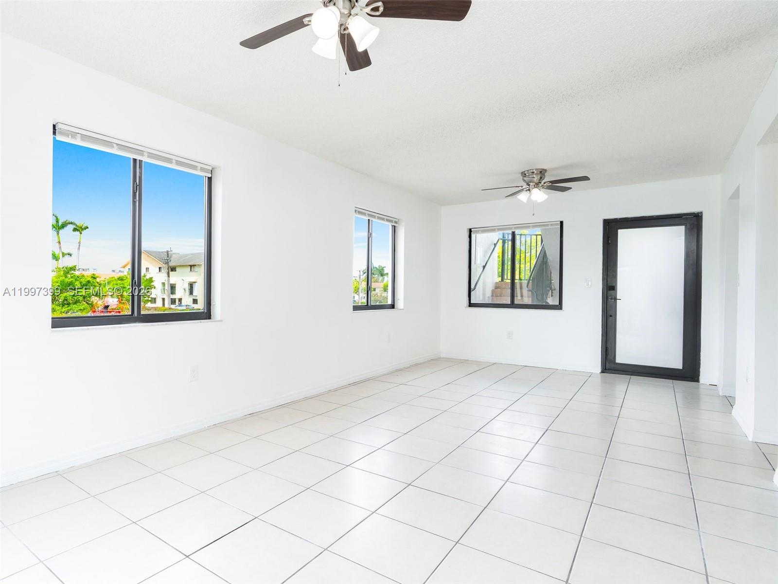 An empty room features white walls, light-colored tile flooring, two ceiling fans, and several windows — 1490 W 42nd Pl # 206, Hialeah FL 33012, HIALEAH, FL