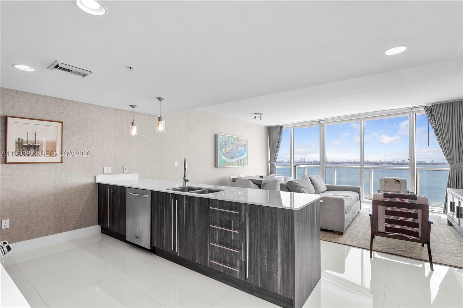 A modern kitchen with dark wood cabinets and white countertops overlooks a living area and ocean — 665 NE 25th St # 2102, Miami FL 33137, MIAMI, FL