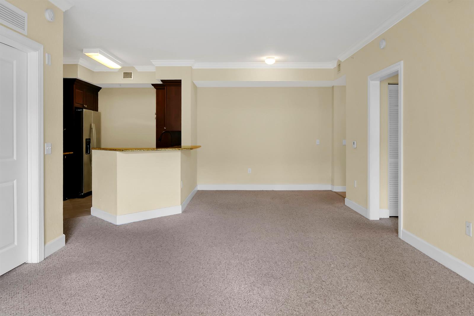 An empty room with beige walls, tan carpeting, and an open kitchen with dark cabinets — 1690 Renaissance Commons Boulevard # 1103, Boynton Beach FL 33426, BOYNTON BEACH, FL