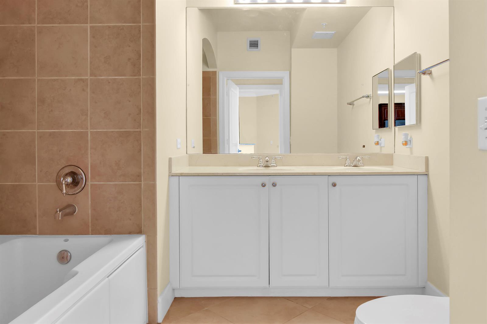 A bathroom features a white double vanity, a large mirror, and a tiled bathtub surround — 1690 Renaissance Commons Boulevard # 1103, Boynton Beach FL 33426, BOYNTON BEACH, FL