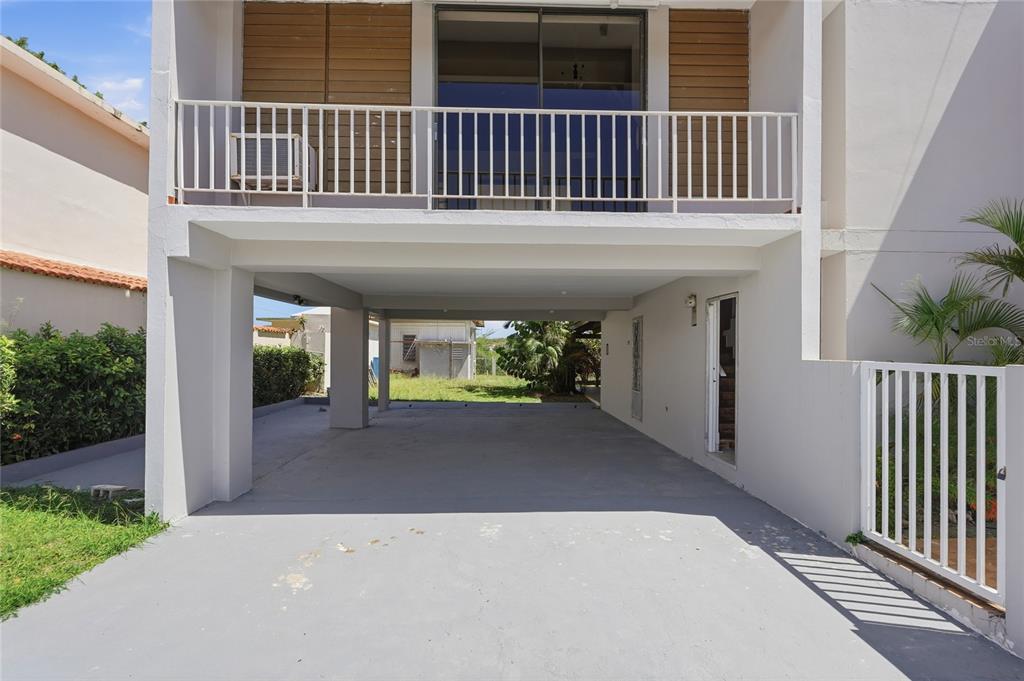 A ground-level concrete carport sits beneath a second-story balcony with a white metal railing — Playa Santa CALLE 3 UNIT D-13, ENSENADA, PR