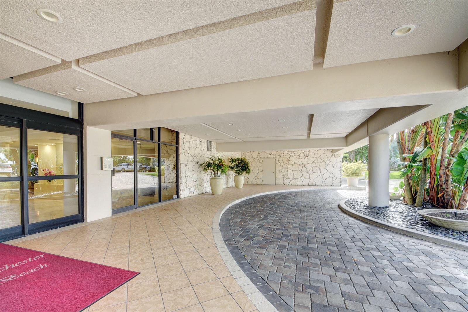 A covered building entrance features a stone wall, tiled floor, and a paved circular driveway — 3250 S Ocean Boulevard # 207s, Palm Beach FL 33480, PALM BEACH, FL
