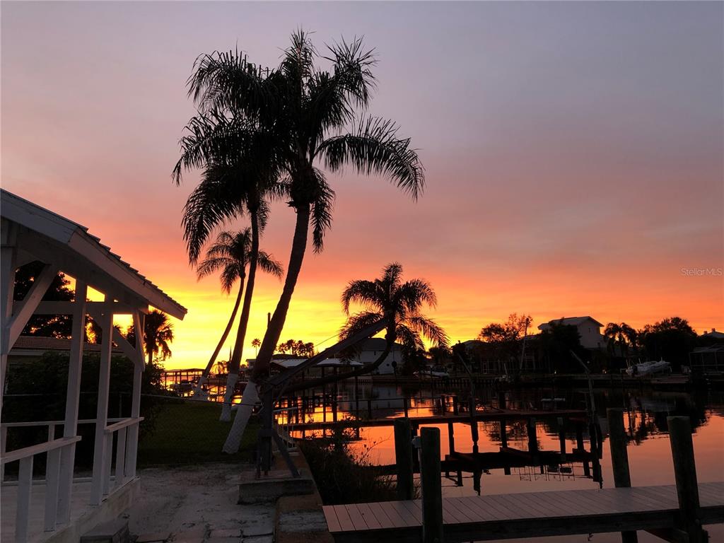 A vibrant sunset glows behind palm trees overlooking a canal with a wooden dock — 2395 HANOVER DR, DUNEDIN, FL