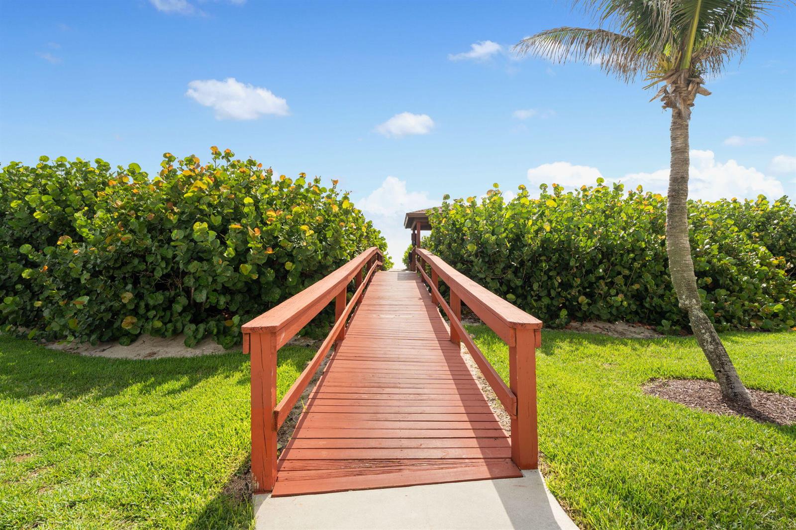 A wooden footbridge leads through lush green bushes toward a beach area under blue skies — 7420 S Ocean Drive # 314, Jensen Beach FL 34957, JENSEN BEACH, FL