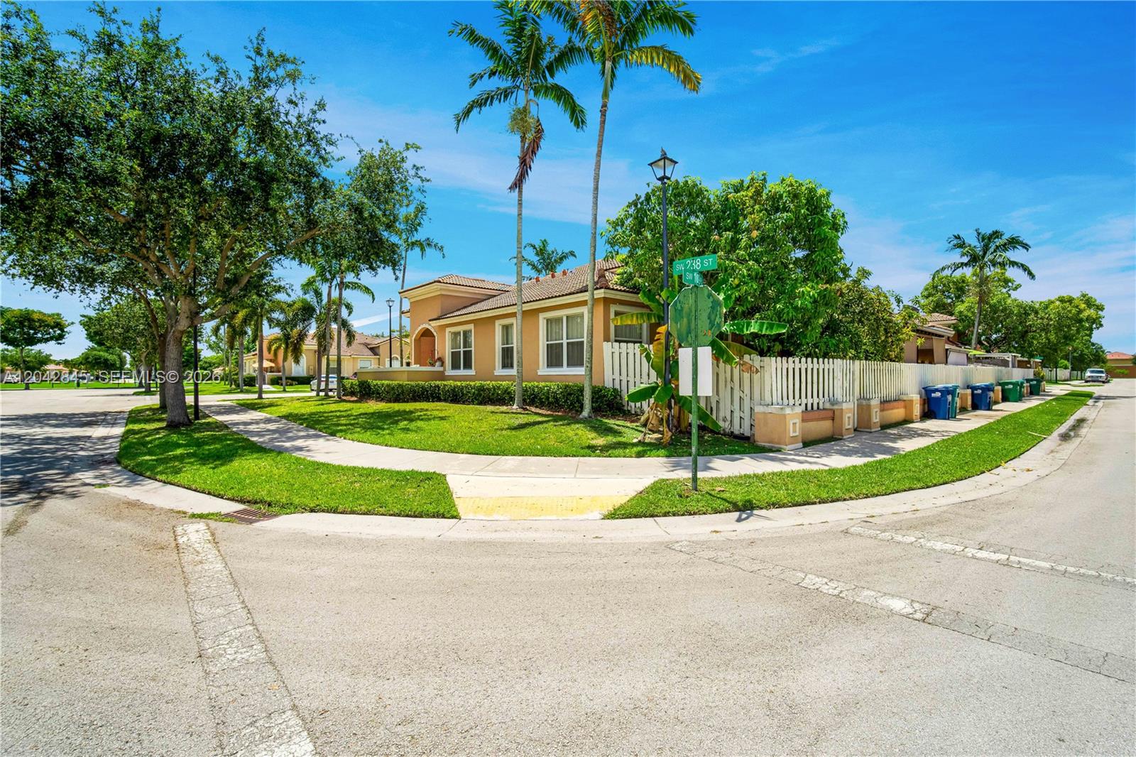 A tan house with a white fence sits on a corner lot with palm trees — 23801 SW 109th Pl, Homestead FL 33032, HOMESTEAD, FL