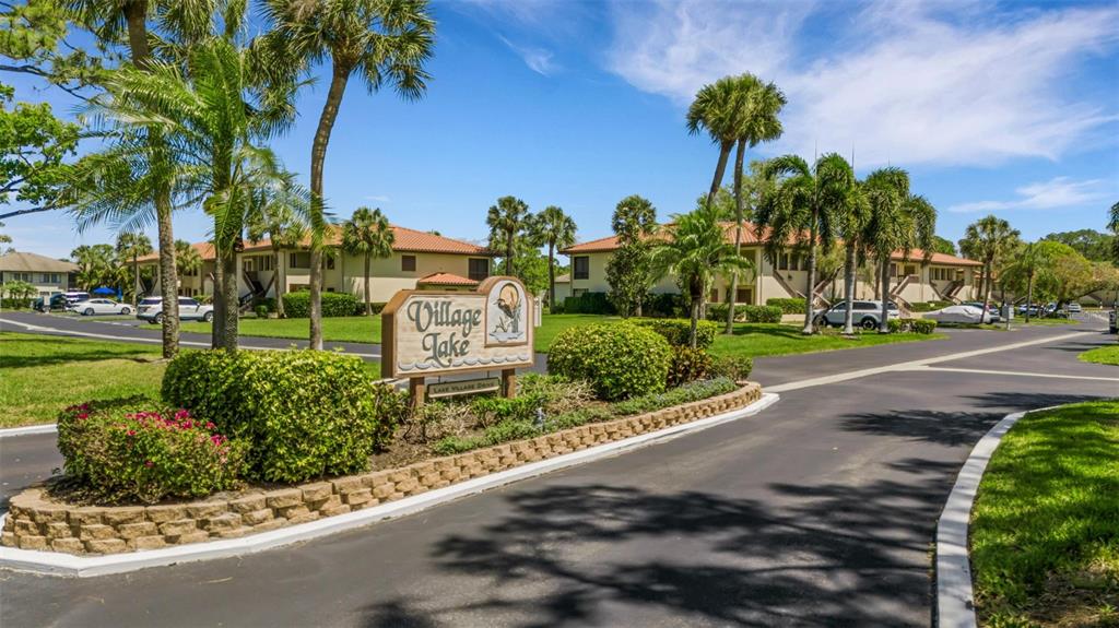 A sign for Village Lake stands at the entrance of a residential complex with palm trees — 5252 LAKE VILLAGE DRIVE UNIT 54, SARASOTA, FL
