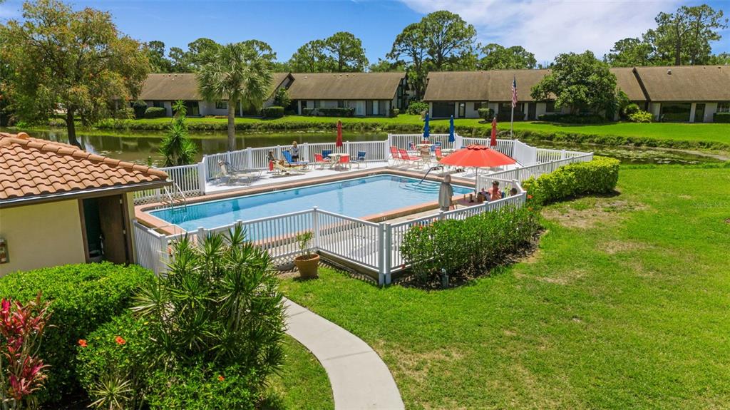 An outdoor swimming pool with a white fence is surrounded by a lawn and buildings — 5252 LAKE VILLAGE DRIVE UNIT 54, SARASOTA, FL