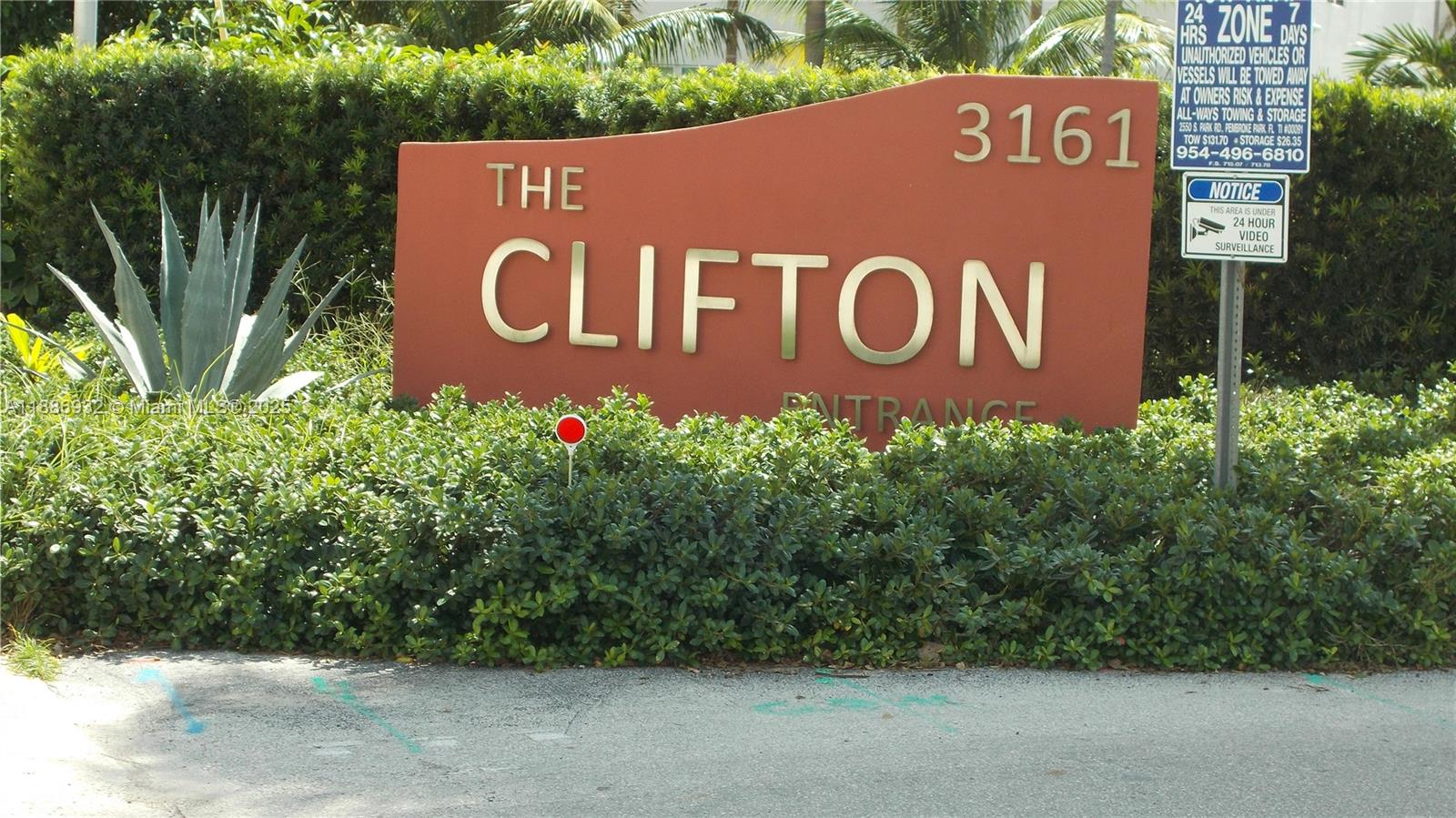 A brown monument sign for The Clifton stands behind a hedge with surveillance signage — 3161 S Ocean Dr # 505, Hallandale Beach FL 33009, HALLANDALE BEACH, FL
