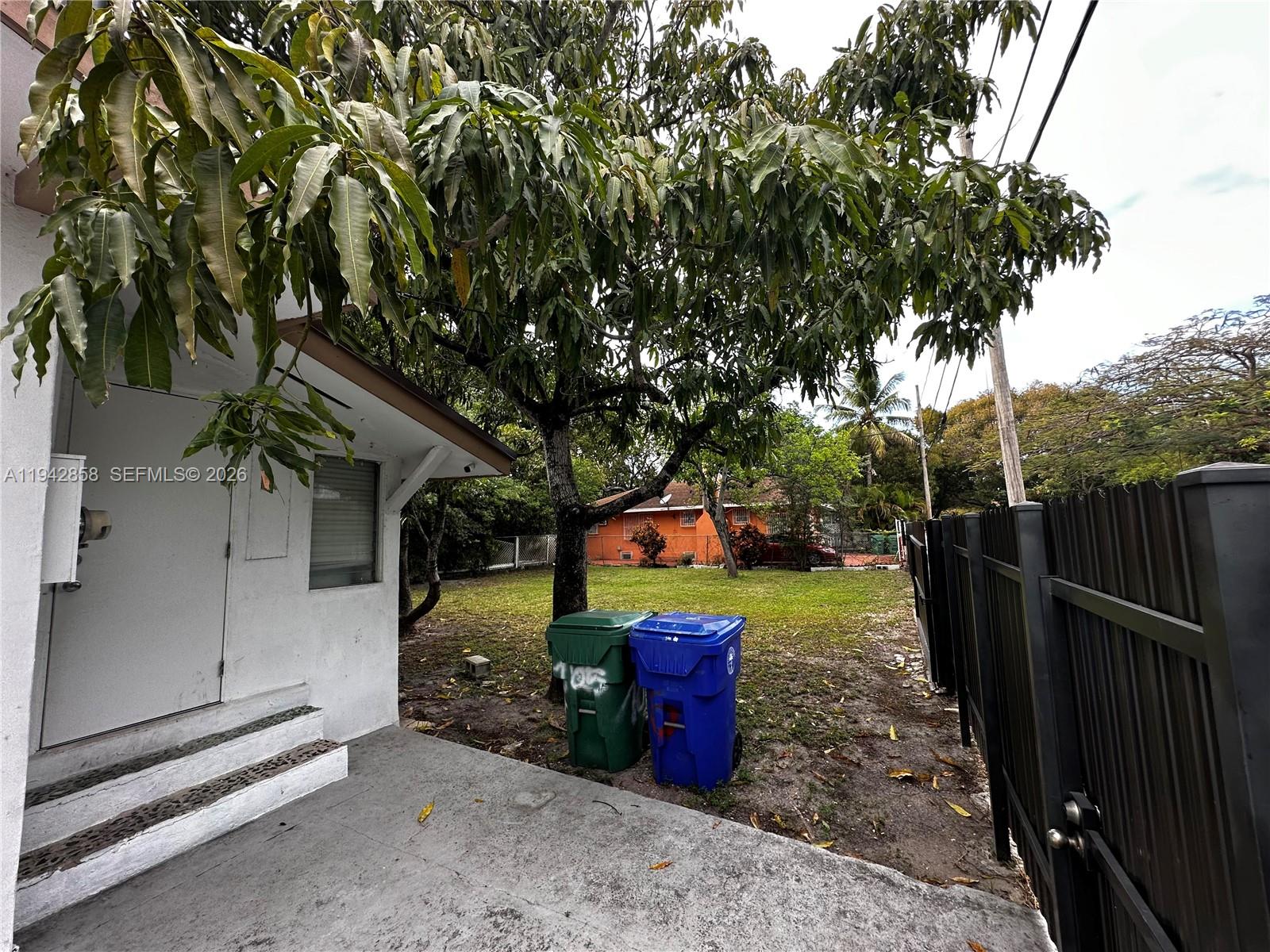 A concrete patio area features two trash bins and a large tree beside a building — 105 NW 31st St # 1, Miami FL 33127, MIAMI, FL