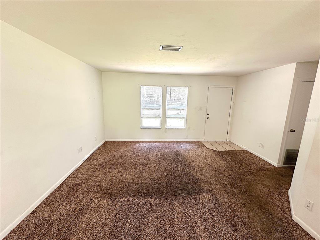 An empty living room features brown wall-to-wall carpeting, white walls, two windows, and a door — 4146 NW 30TH TERRACE, GAINESVILLE, FL