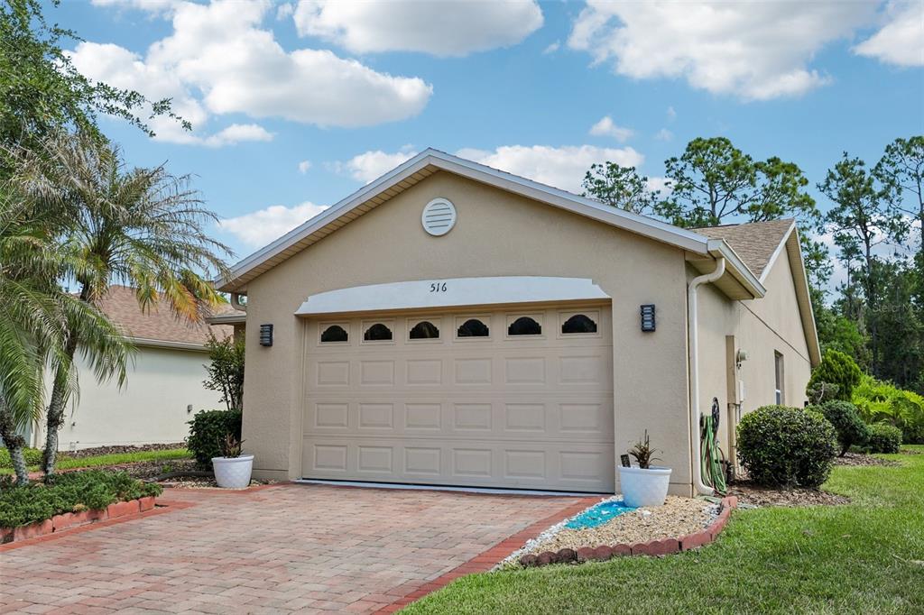 A beige house exterior features a two-car garage, a brick paver driveway, and a palm tree — 516 MILLIFIORA LANE, POINCIANA, FL
