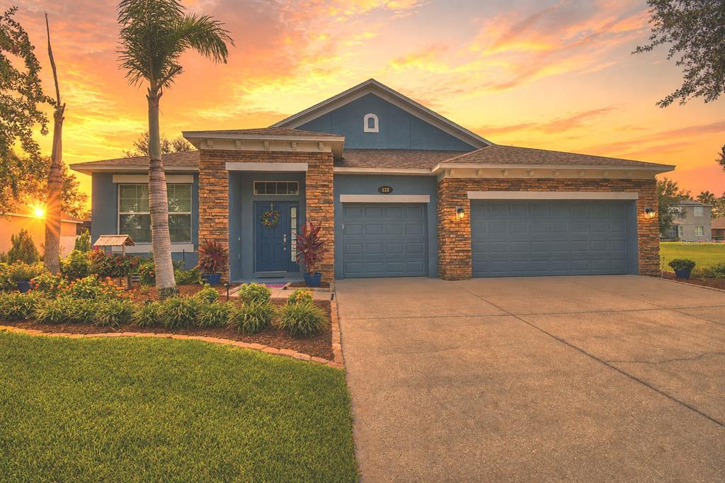 A blue single-story house with a stone facade, a three-car garage, and a sunset sky — 112 PECORRI COURT, AUBURNDALE, FL