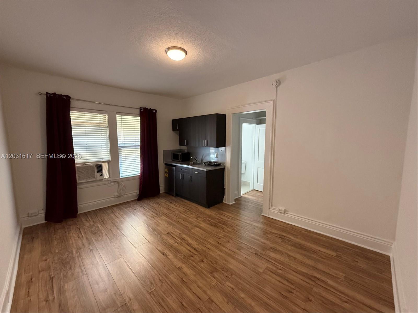 A small studio apartment features dark kitchen cabinets, wood-look flooring, and a window with curtains — 126 Mendoza Ave # 2A, Coral Gables FL 33134, CORAL GABLES, FL