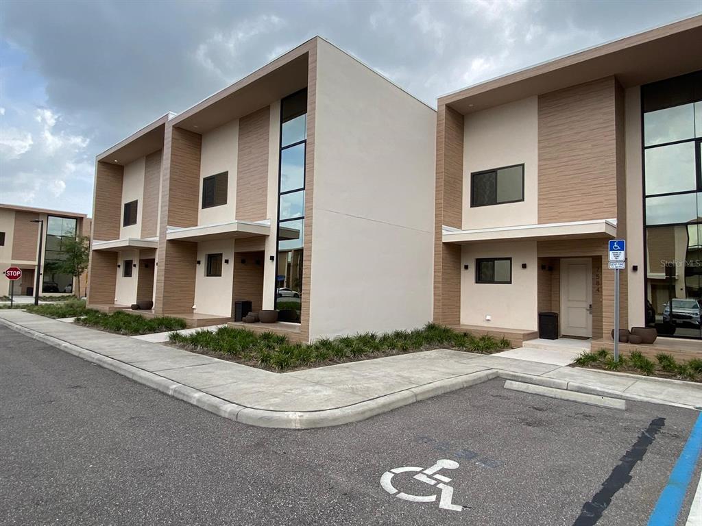 A modern two-story townhouse exterior features light-colored stucco, wood-look siding, and a designated accessible parking space — 7582 BROOKLYN DRIVE, KISSIMMEE, FL