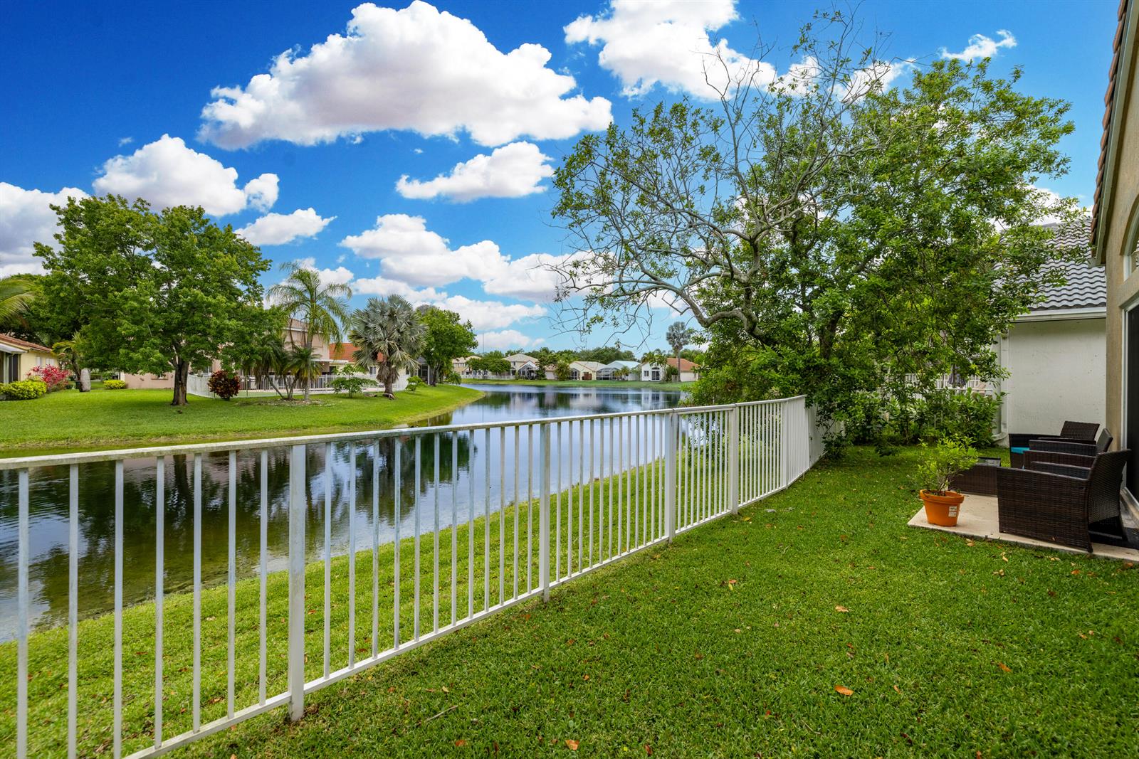 A white metal fence borders a grassy backyard overlooking a lake with trees and houses — 6513 Sandpiper Drive, Coconut Creek FL 33073, COCONUT CREEK, FL