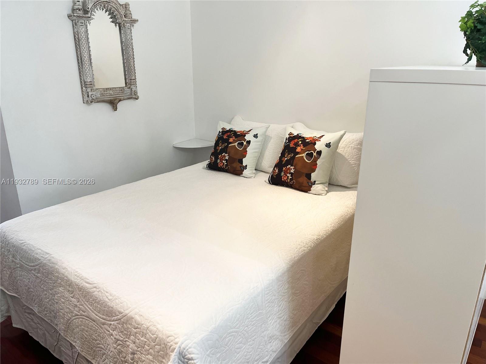 A bedroom features a white bed with decorative pillows, a wall-mounted mirror, and a shelf — 1340 Drexel Ave # 406, Miami Beach FL 33139, MIAMI BEACH, FL
