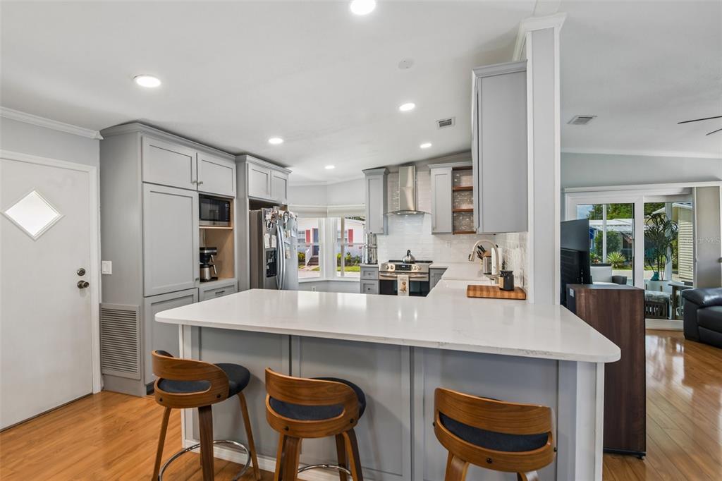 A kitchen features gray cabinetry, white countertops, a breakfast bar with three stools, and wood flooring — 276 PELICAN DRIVE N, OLDSMAR, FL