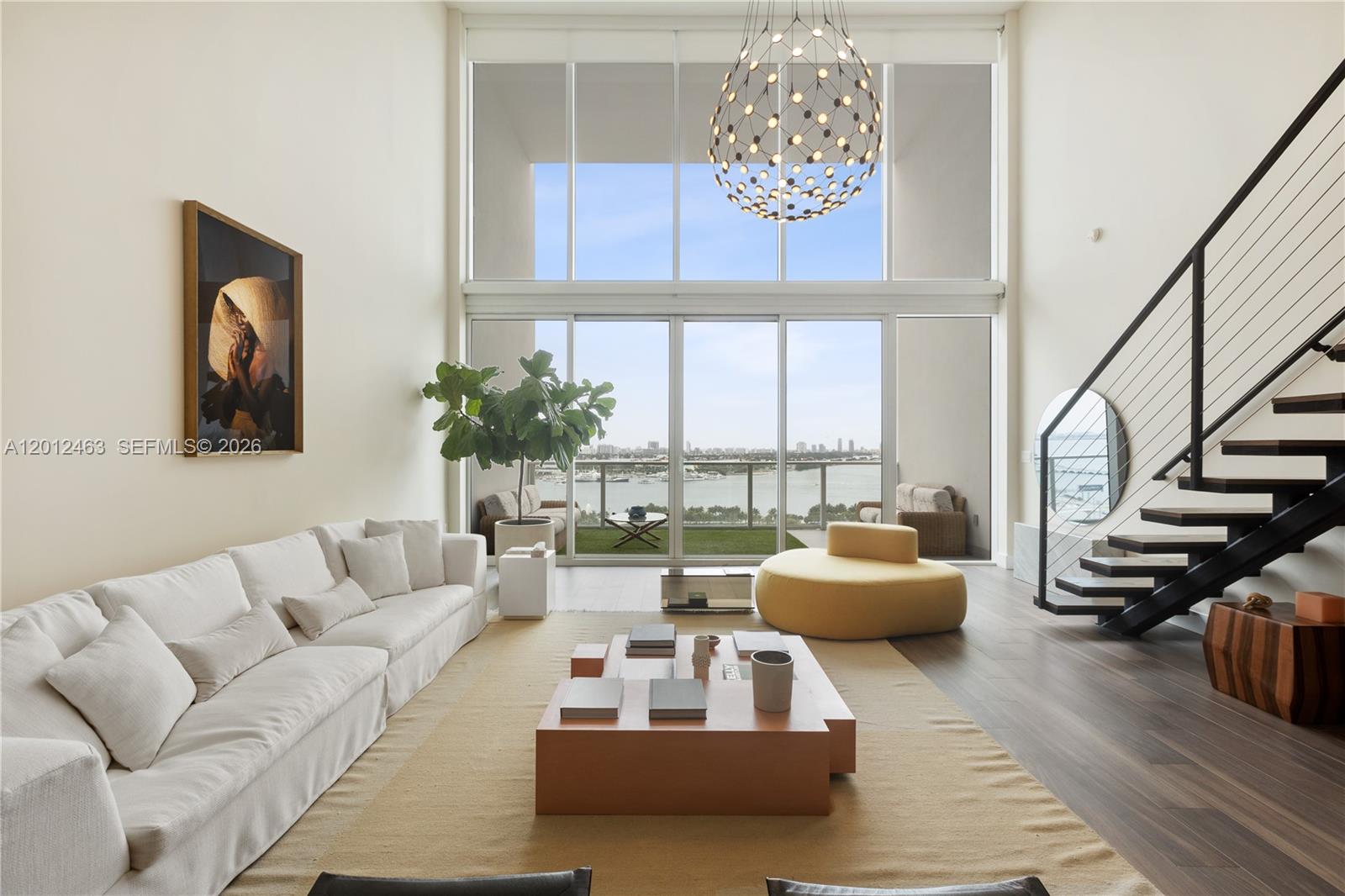 A modern living room features double-height windows, a floating staircase, and a view of water — 1040 Biscayne Blvd # 1602, Miami FL 33132, MIAMI, FL