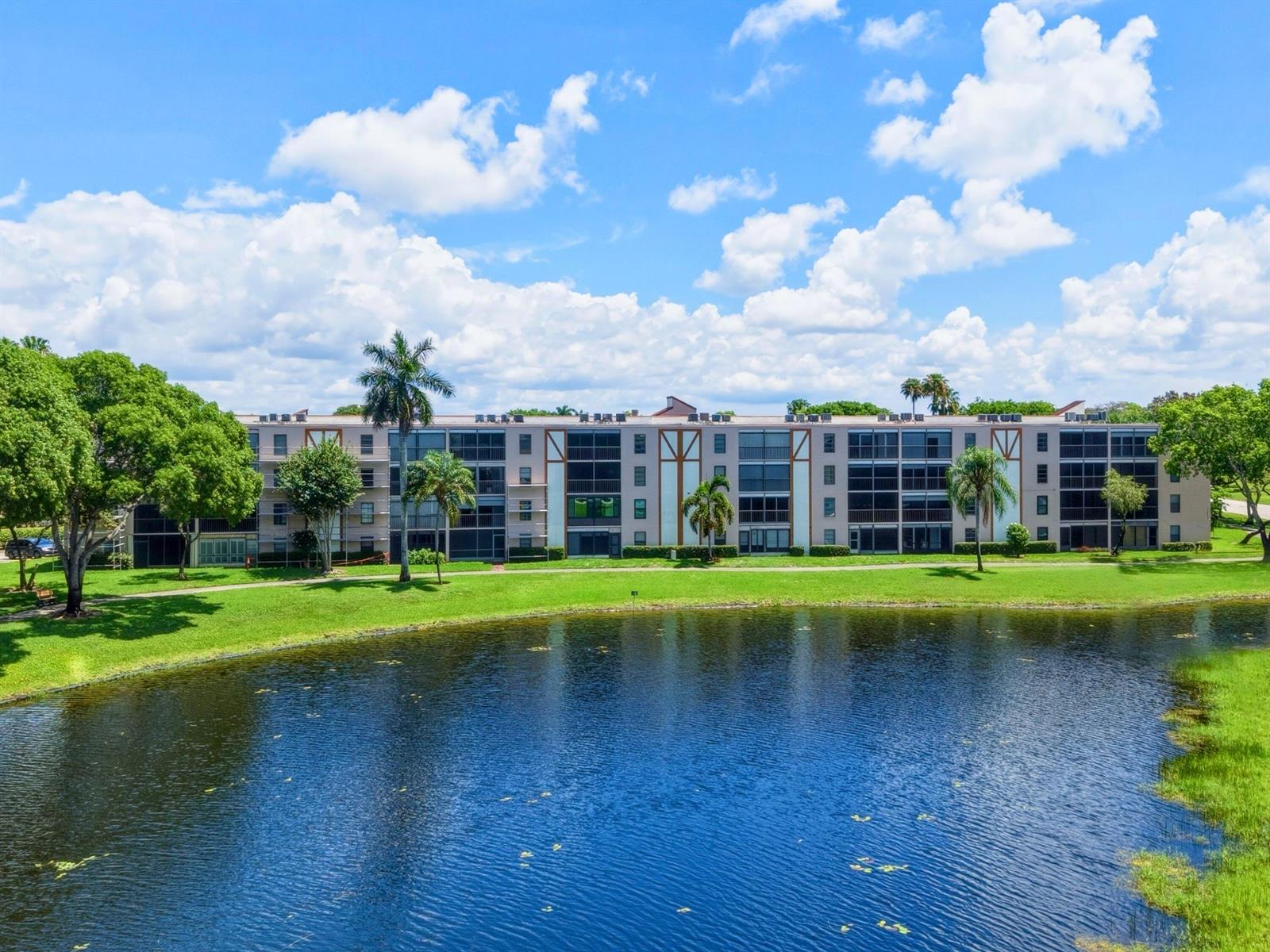 A multi-story apartment building sits behind a pond with palm trees and green landscaping — 7280 Amberly Lane # 201, Delray Beach FL 33446, DELRAY BEACH, FL