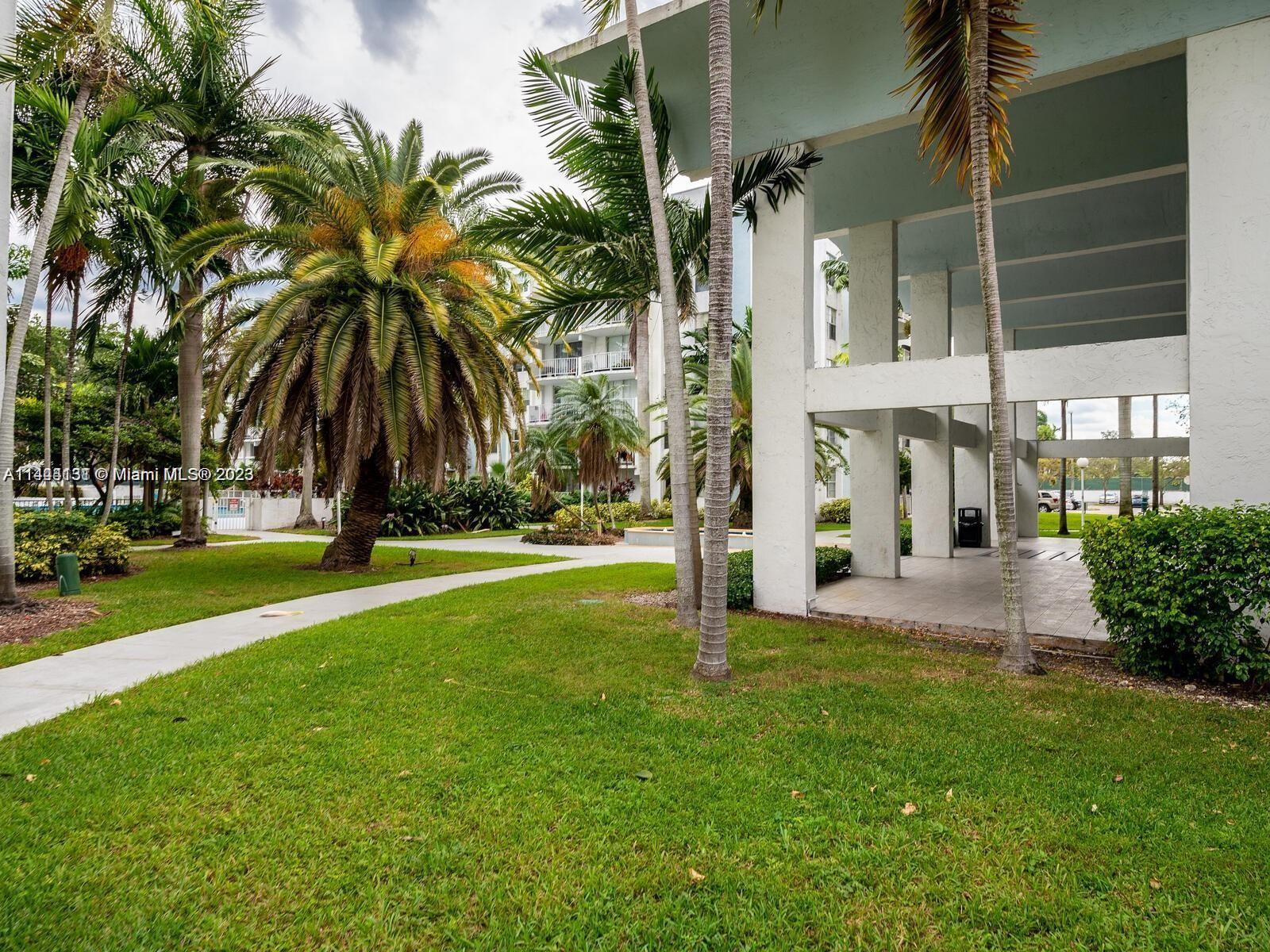 A paved walkway winds through a grassy courtyard with palm trees and a covered building entrance — 496 NW 165th St Rd # D112, Miami FL 33169, MIAMI, FL