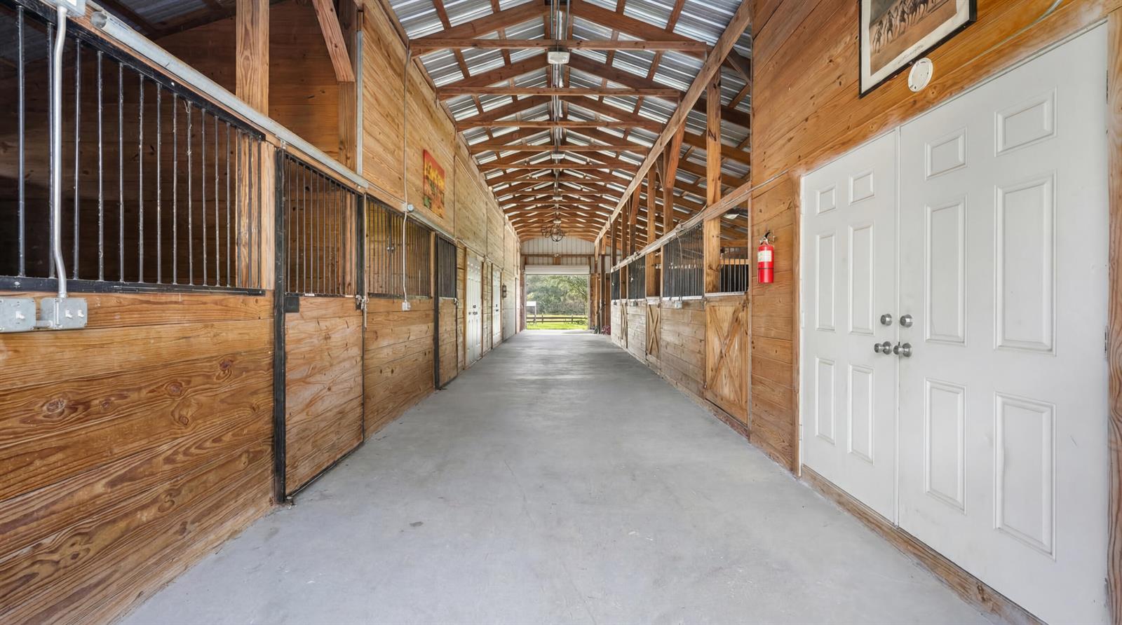 A long, concrete-floored horse barn aisle features wooden stalls with metal bars on both sides — 12440 Tangerine Boulevard, FL 33412, FL