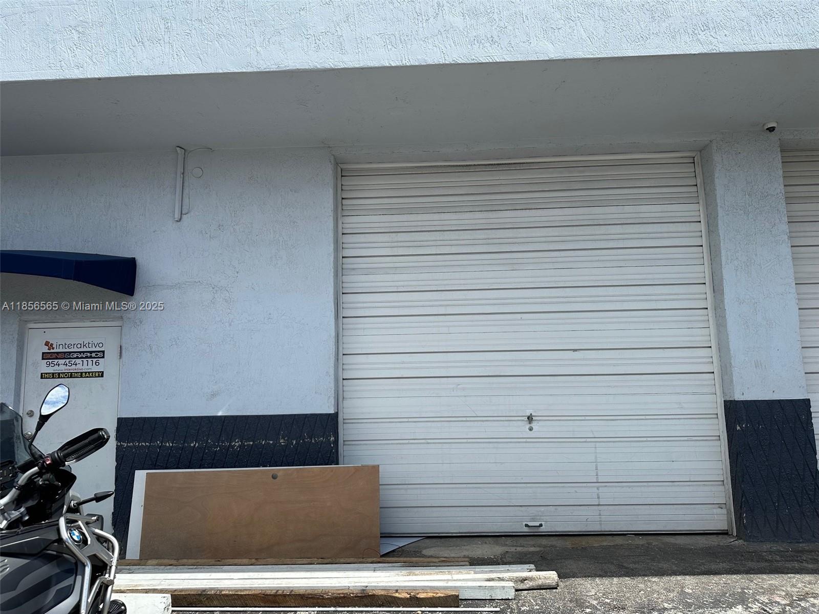 A white exterior wall features a large roll-up metal door and a side entry door — 470 Ansin Blvd # 470H, Hallandale Beach FL 33009, HALLANDALE BEACH, FL