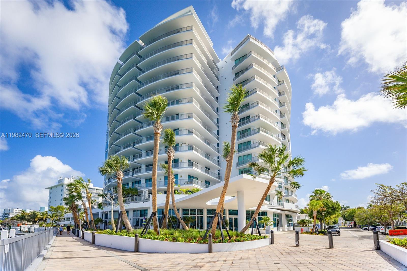 A modern multi-story residential building with curved balconies stands behind palm trees and a walkway — 1445 16th St # 1101, Miami Beach FL 33139, MIAMI BEACH, FL