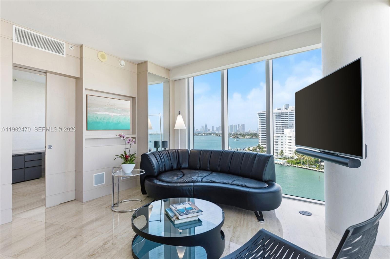 A living room features a curved black leather sofa, glass coffee table, and waterfront views — 1445 16th St # 1101, Miami Beach FL 33139, MIAMI BEACH, FL
