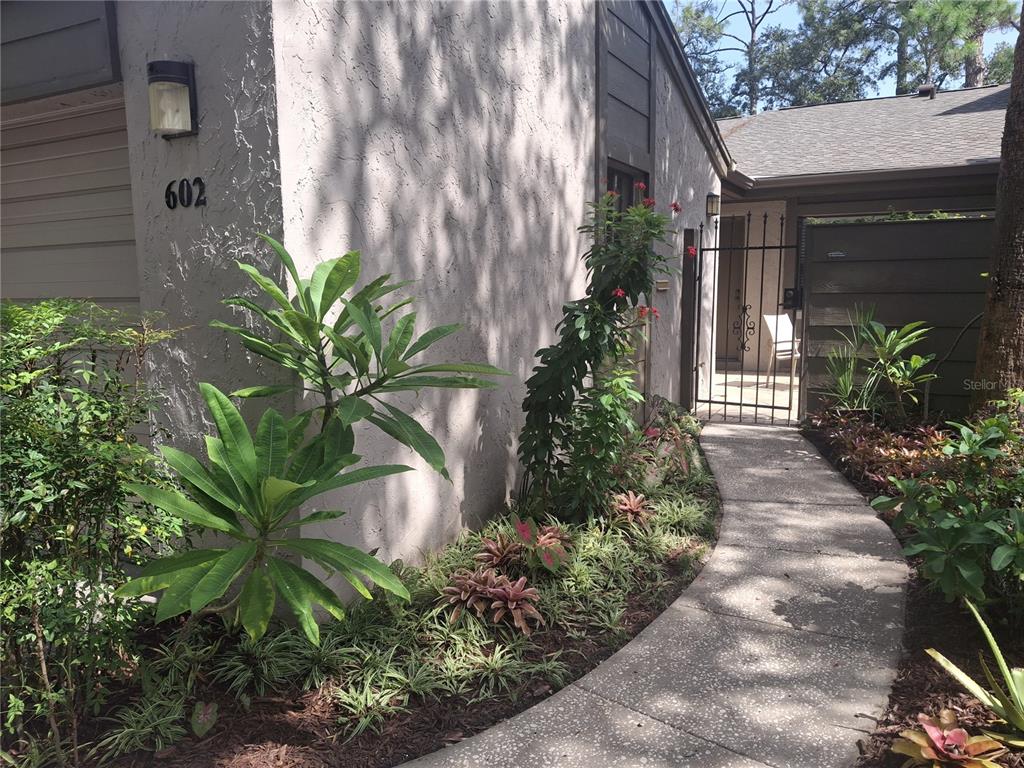 A concrete walkway leads to a gated entrance beside a stucco building with landscaping — 602 WOODRIDGE DRIVE, CASSELBERRY, FL
