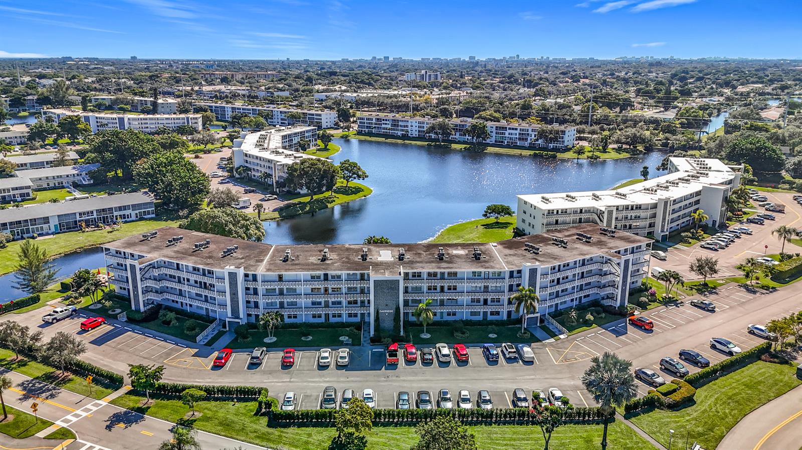 An aerial view shows multi-story apartment buildings surrounding a lake with a large parking lot — 2114 Oakridge V, Deerfield Beach FL 33442, DEERFIELD BEACH, FL