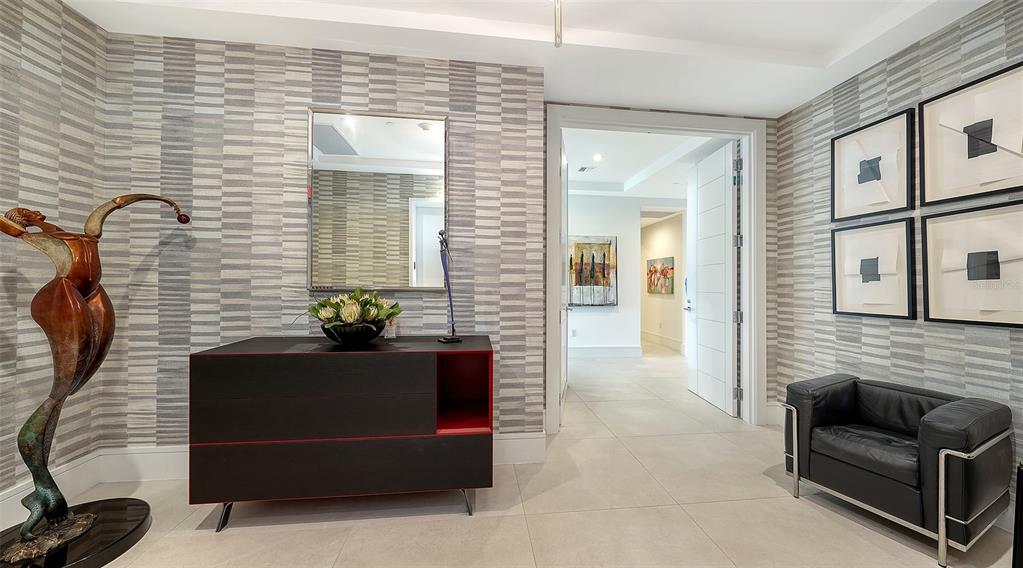 A modern entryway features patterned wallpaper, a dark console table, a sculpture, and a leather chair — 401 QUAY COMMONS UNIT 1004, SARASOTA, FL