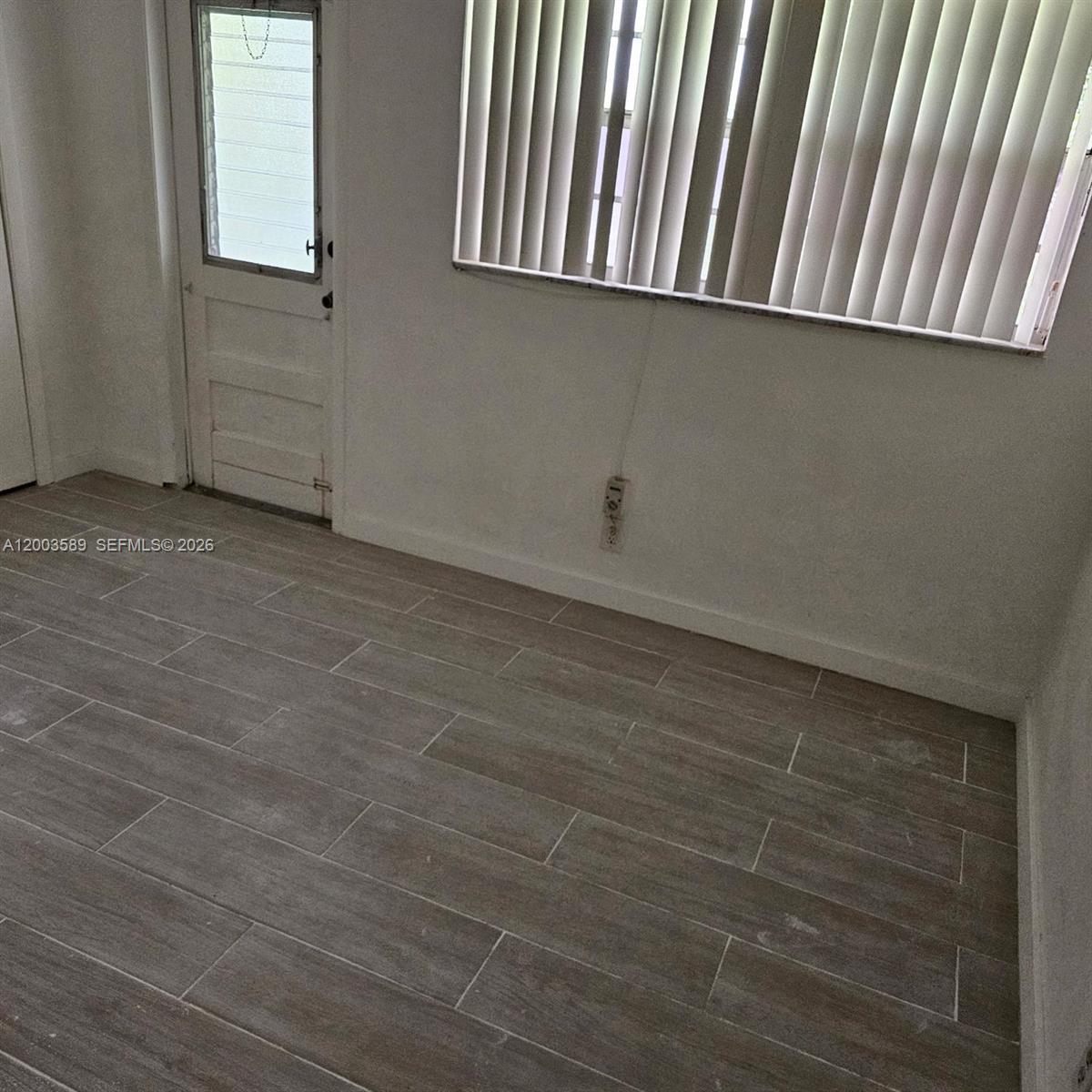 An empty room features light-colored wood-look tile flooring, white walls, vertical blinds, and a door — 230 SW 11th Ave # 14, Hallandale Beach FL 33009, HALLANDALE BEACH, FL
