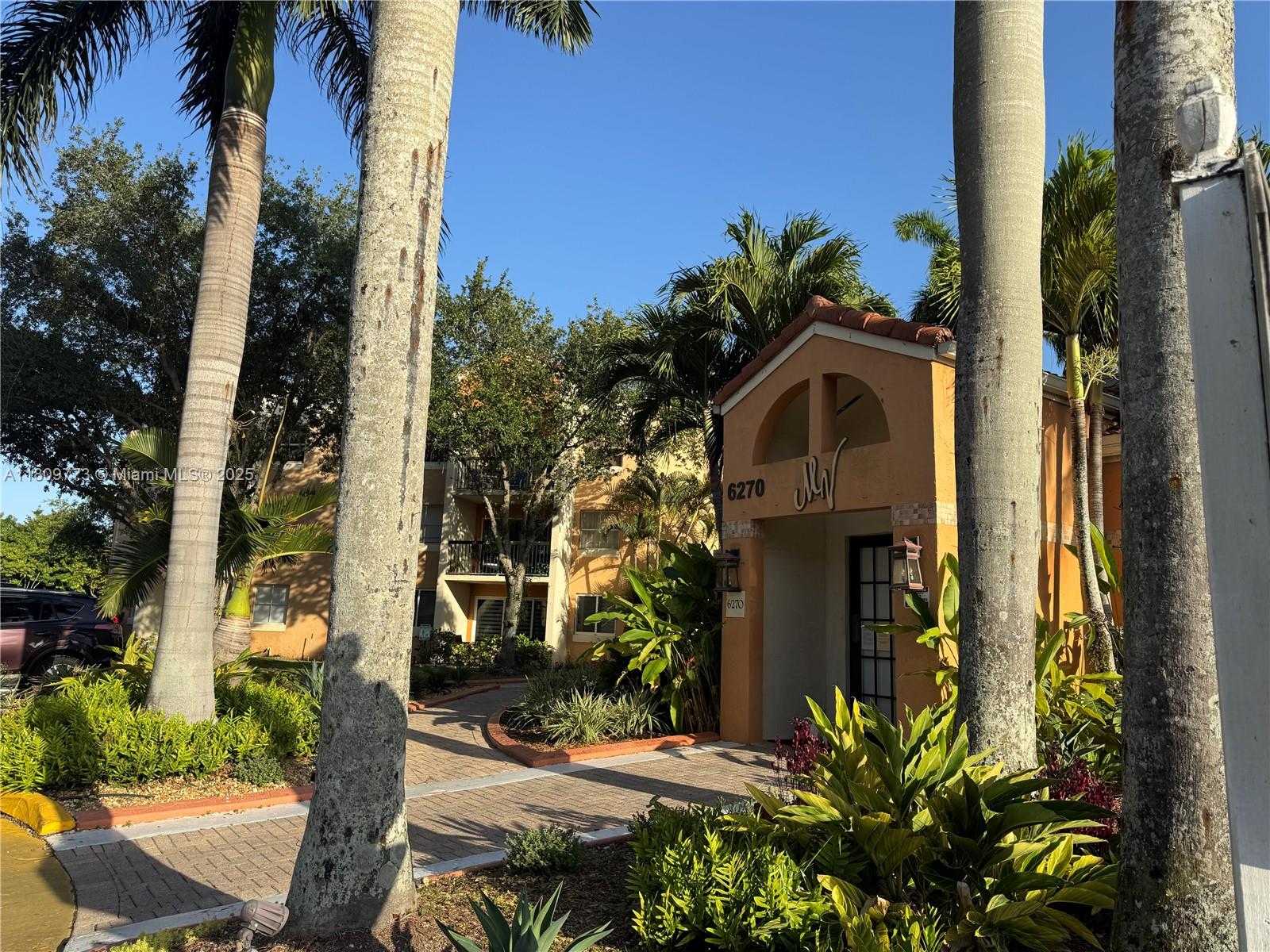 A paved walkway leads to the entrance of a multi-story apartment building with palm trees — 6276 NW 186th St # 305, Hialeah FL 33015, HIALEAH, FL