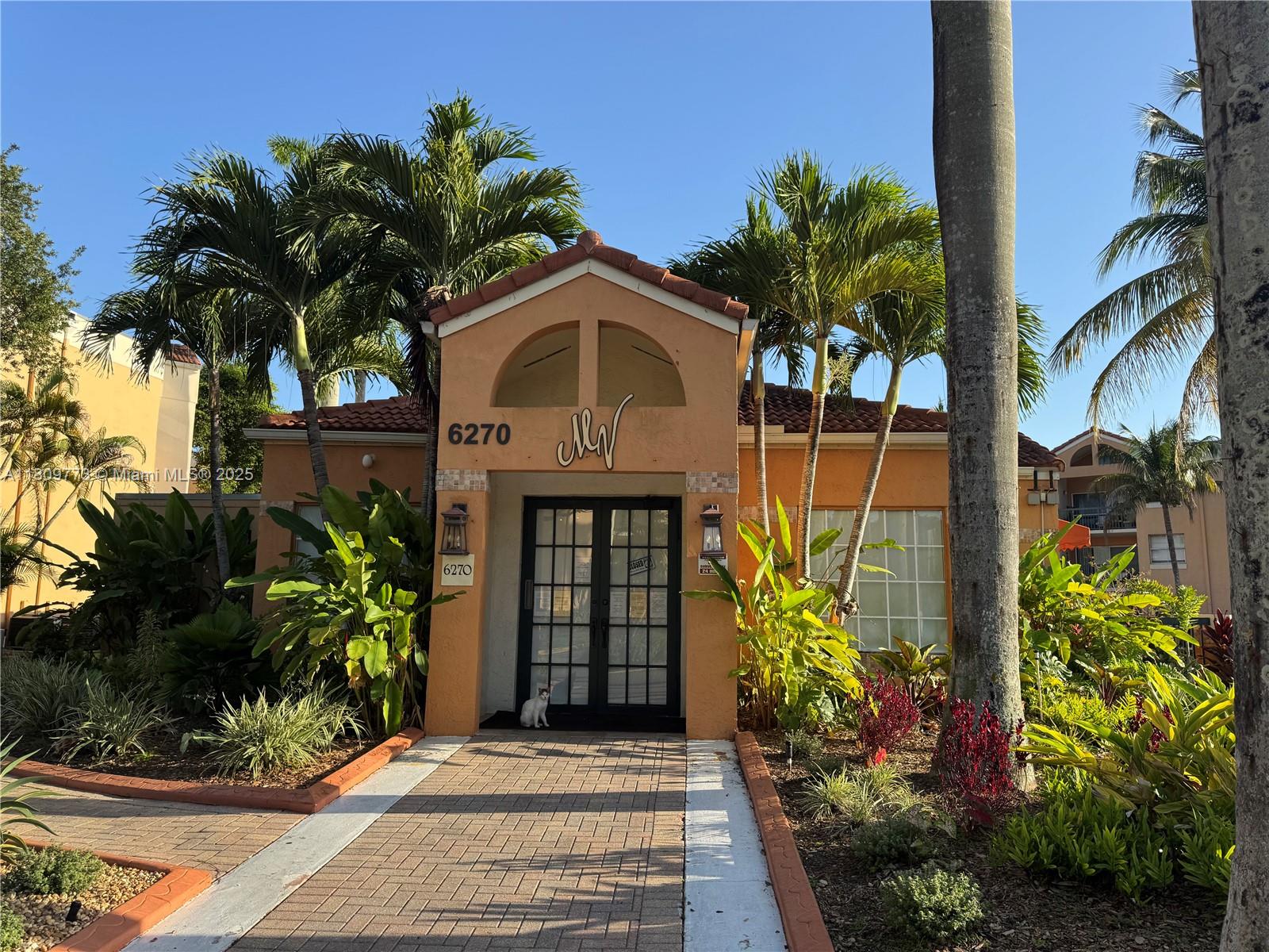 A peach-colored building entrance features a tiled walkway, palm trees, and a small cat — 6276 NW 186th St # 305, Hialeah FL 33015, HIALEAH, FL