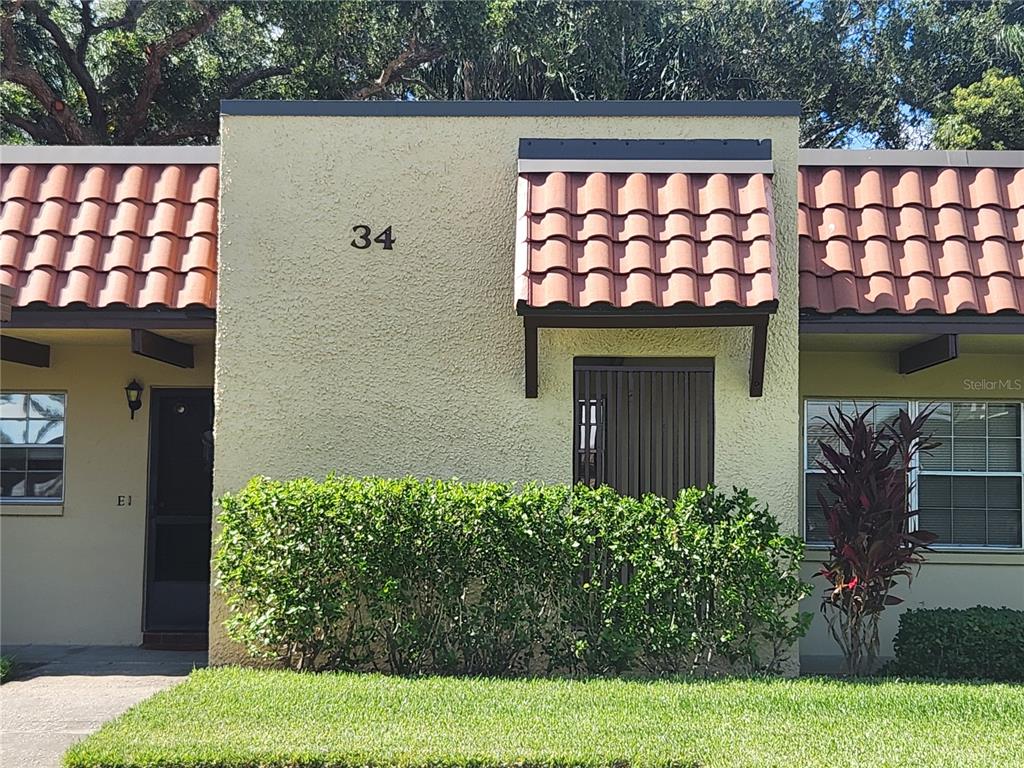 A single-story building exterior features a stucco facade, red tile accents, and a front hedge — 1701 PINEHURST ROAD UNIT 34D, DUNEDIN, FL